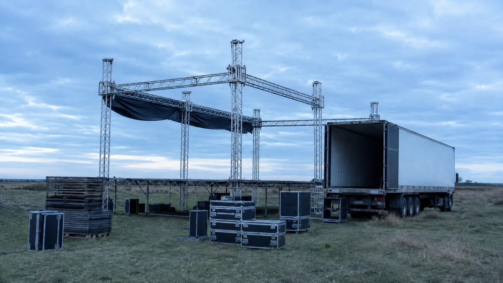 Escenario de evento a medio armar en un campo al amanecer, con estructura de truss, cajas de equipos y un semirremolque con las puertas abiertas